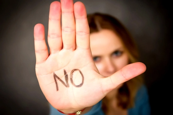 Una mujer joven con una expresión facial de firmeza y desaprobación, levantando la mano en un gesto de "no" o para detener algo, con un fondo desenfocado de un entorno doméstico moderno.